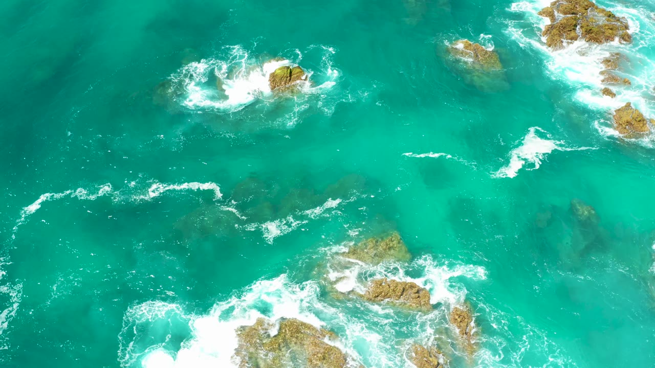 Aerial view at Dreams Los Cabos where teal water surrounds several tan rock clusters and white foam spreads along the lower and right edges, with no visible people.