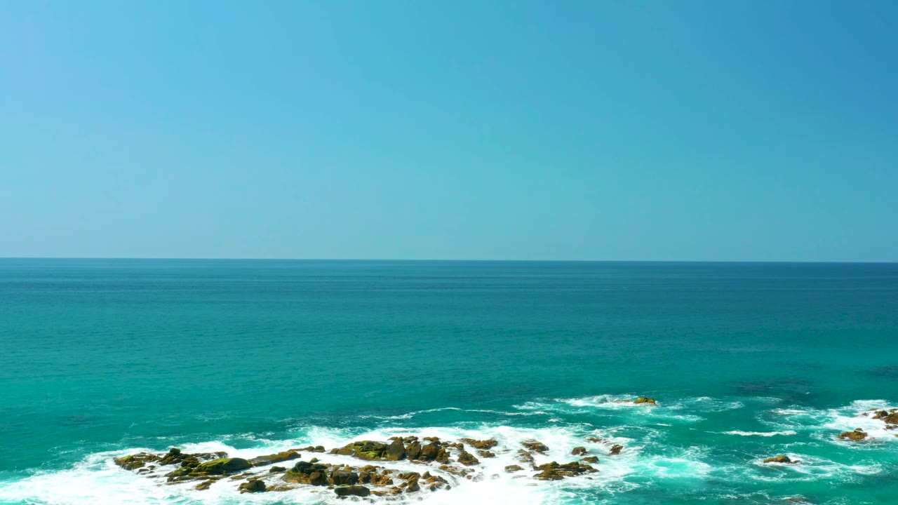 Wide elevated view of turquoise ocean at Dreams Los Cabos with white foam breaking over rocks along the lower edge and no visible people.