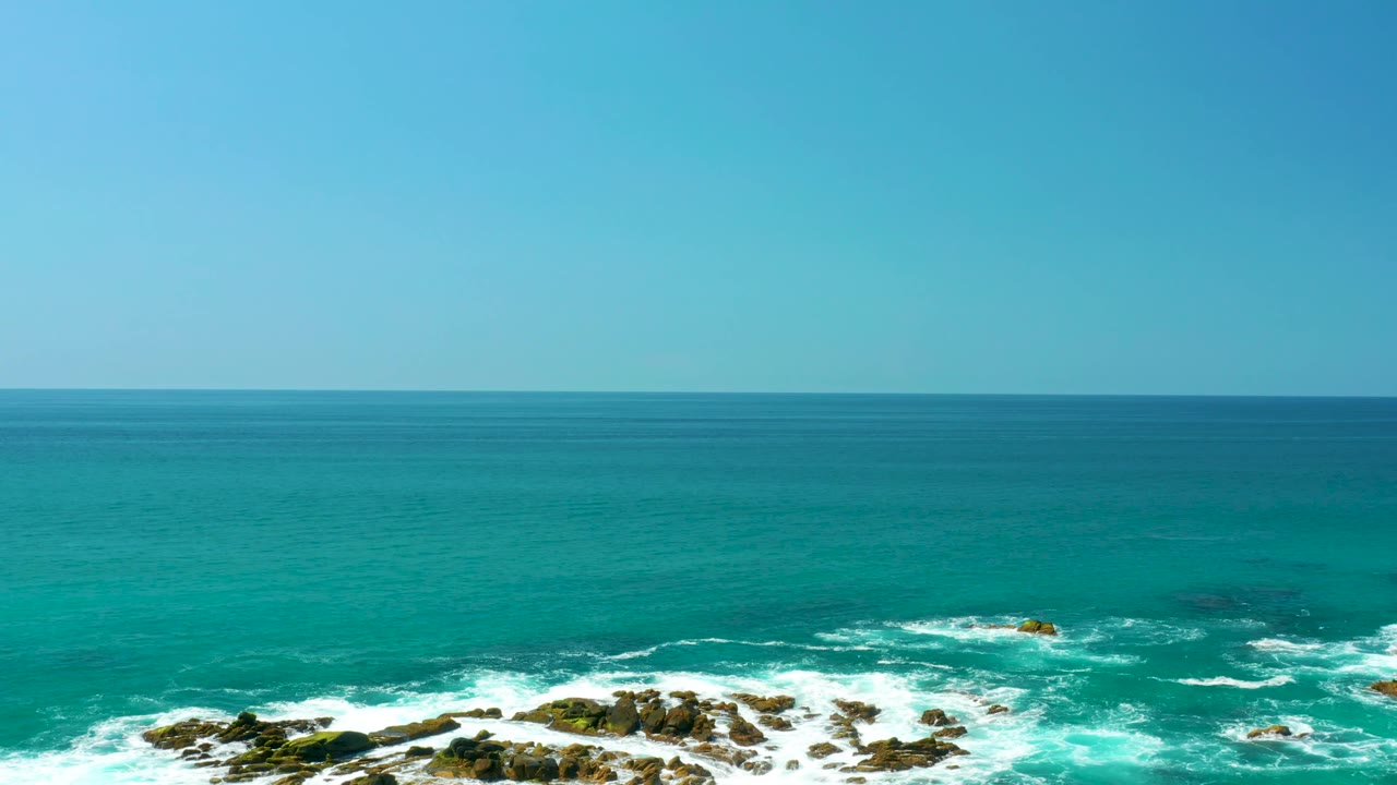 Broad turquoise water fills the Dreams Los Cabos frame as white surf wraps around clustered rocks near the bottom, with no visible people.