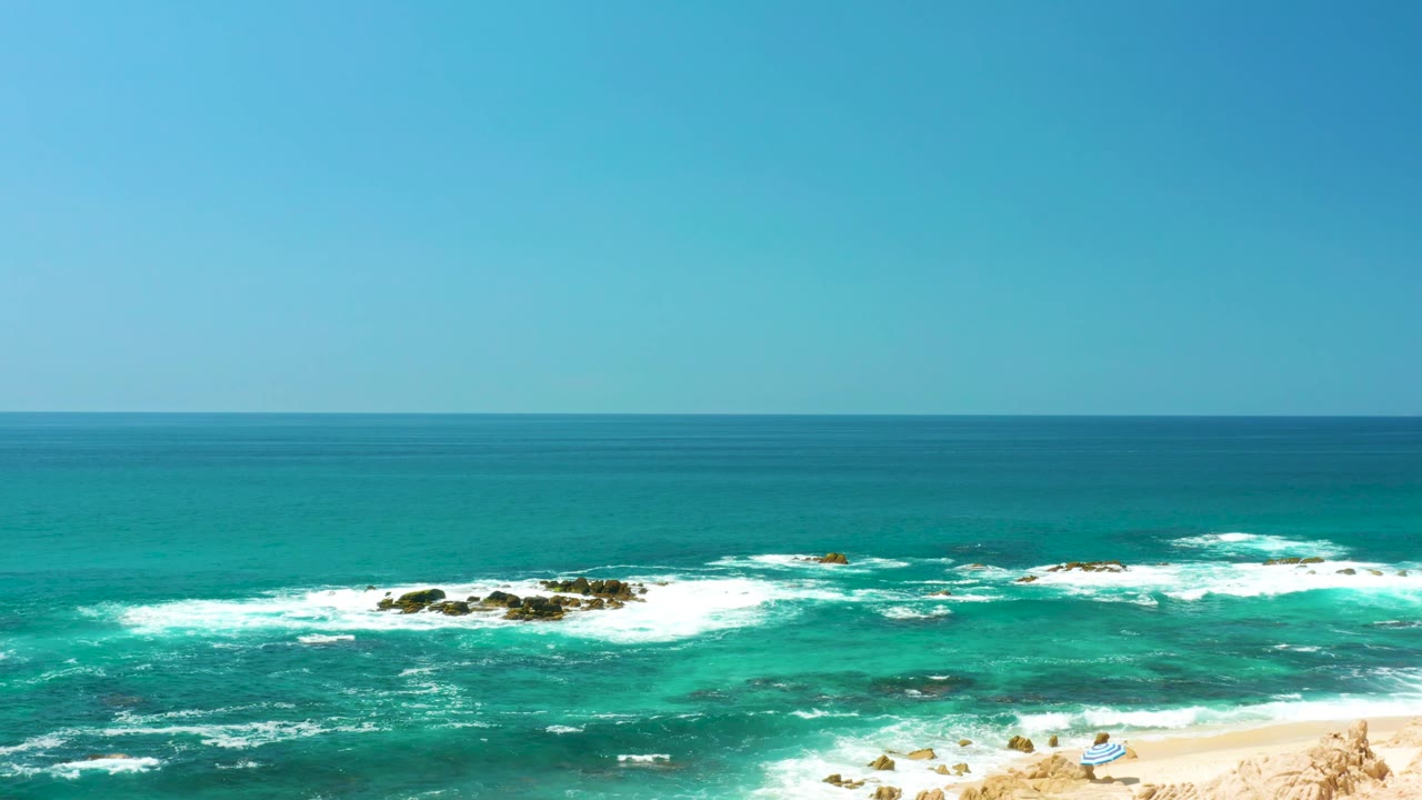 At Dreams Los Cabos in Baja California Sur, the wide frame holds clear sky, turquoise ocean, foaming waves, dark rocks, and a small beach corner with tiny distant beachgoers near the lower edge.