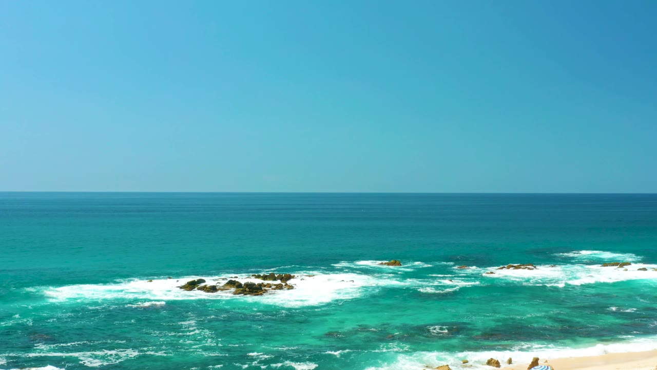 At Dreams Los Cabos in Baja California Sur, white foam spreads across several rocky outcrops in turquoise water beneath a clear blue sky, with only a sliver of beach at the lower edge.