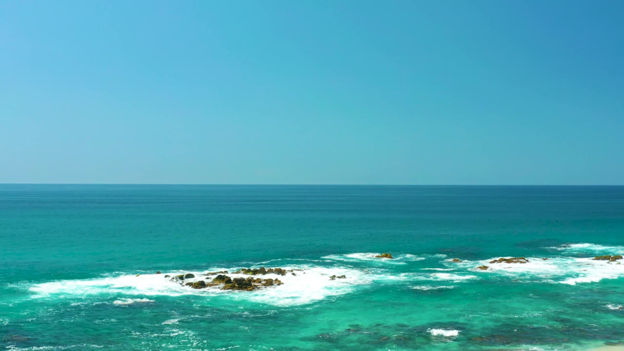 At Dreams Los Cabos in Baja California Sur, the frame shows open turquoise sea, a clean horizon, and white surf breaking over low dark rocks, with no clearly visible people.