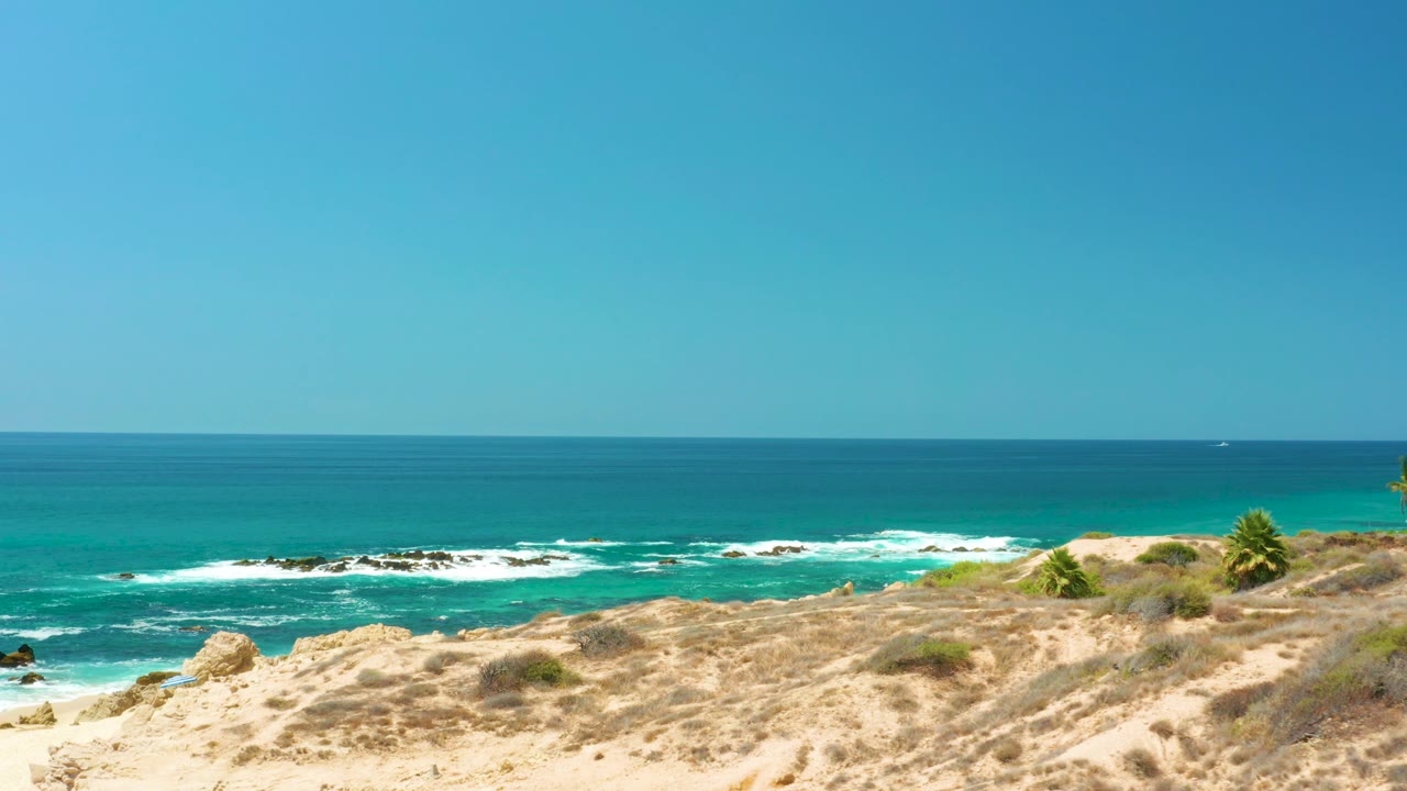 At Dreams Los Cabos in Baja California Sur, a sandy cliff with scrub overlooks turquoise water, white surf, dark rocks, and a clear blue horizon with no visible people.