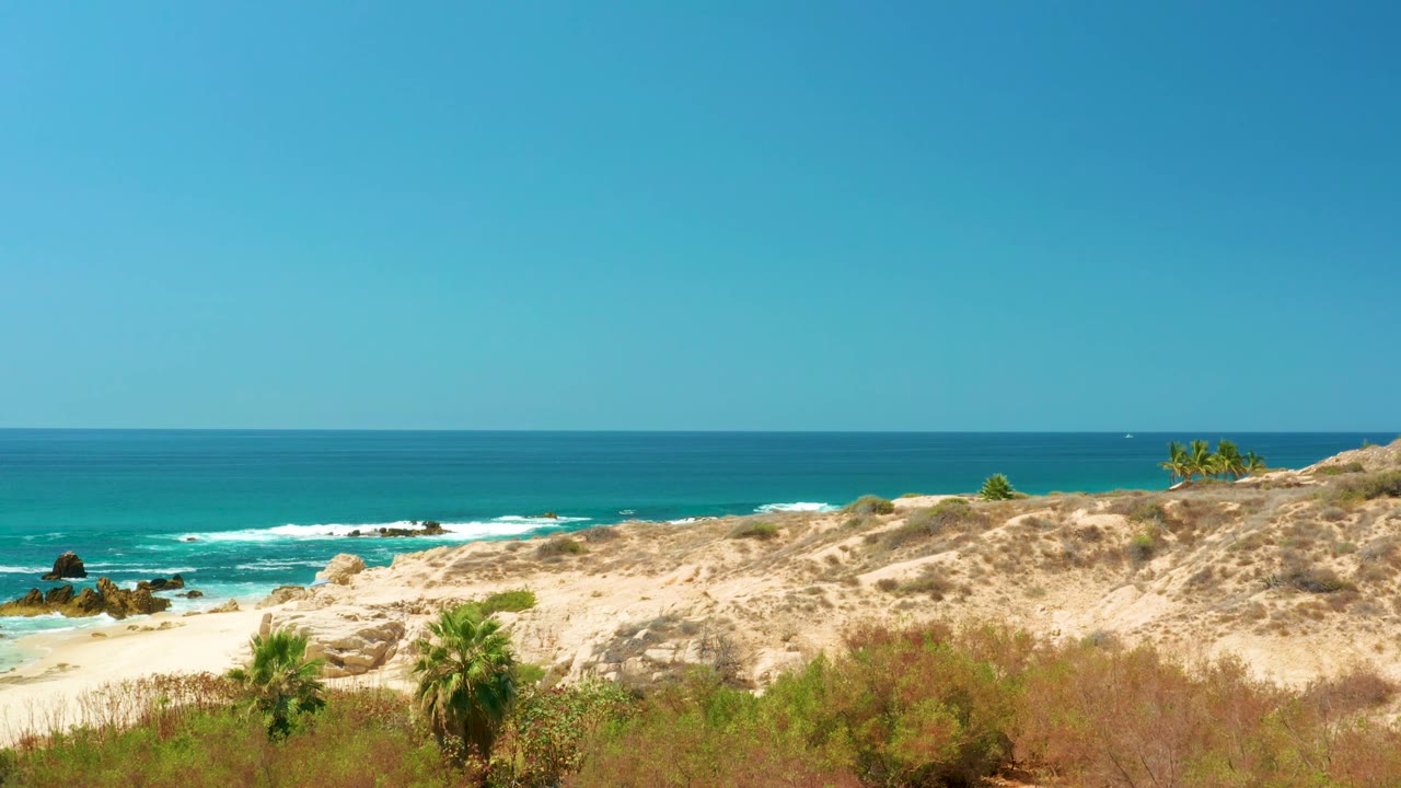 At 0:01, the elevated Dreams Los Cabos shoreline view shows dry dune brush, a sandy beach edge, turquoise water, white waves, rocks, and no visible people.
