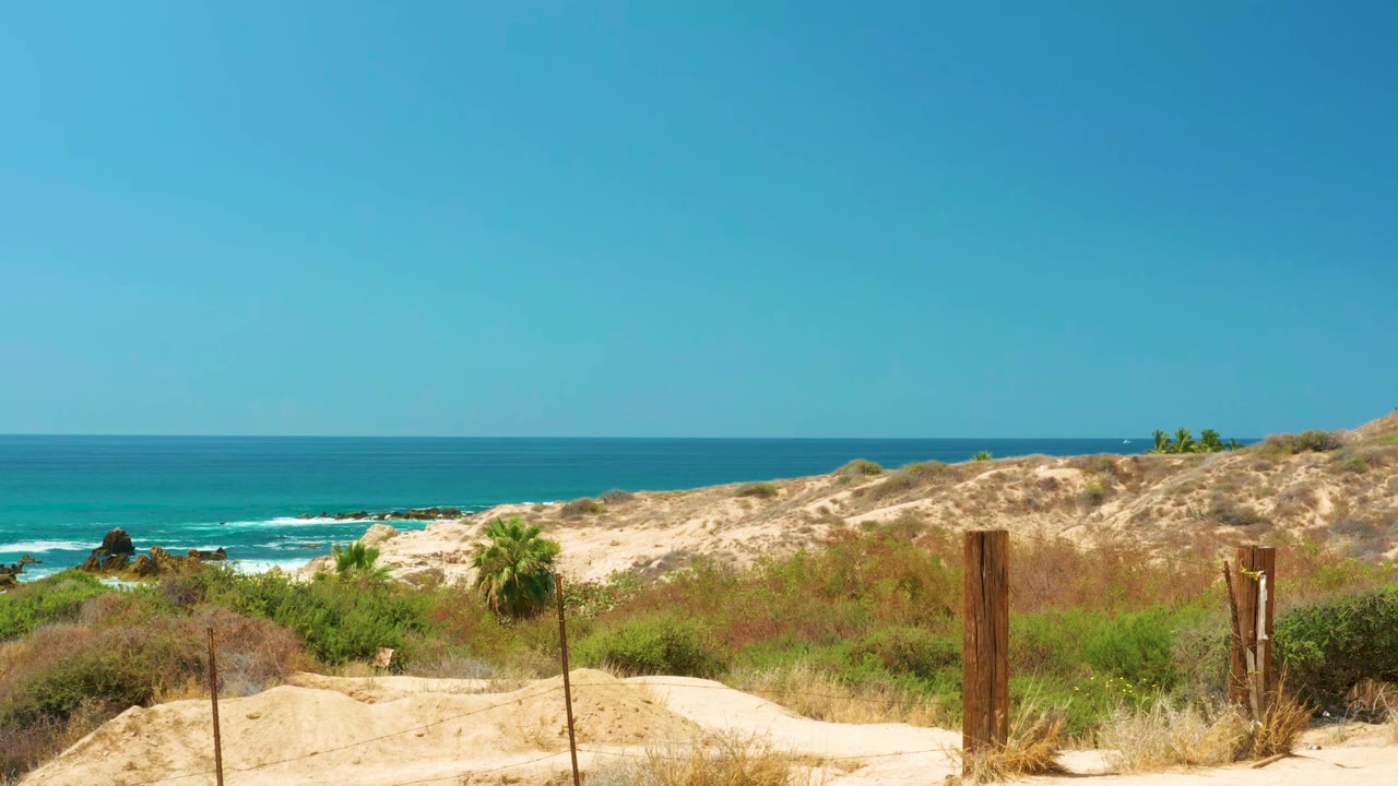 Sandy path, wooden fence posts, dunes, and blue ocean under clear sky.