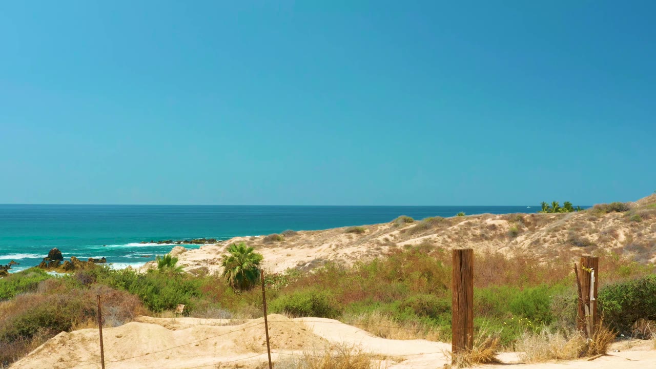 At 0:01, the Dreams Los Cabos dune path remains in the foreground, with fence posts, scrub vegetation, turquoise-blue water, white surf, and no visible people.