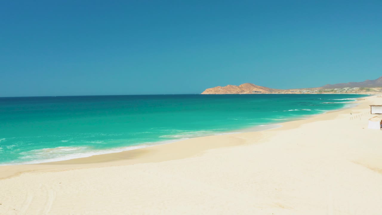 Opening frame of Dreams Los Cabos beach with pale sand in the foreground, turquoise water across the middle, distant hills on the horizon, and no visible people.