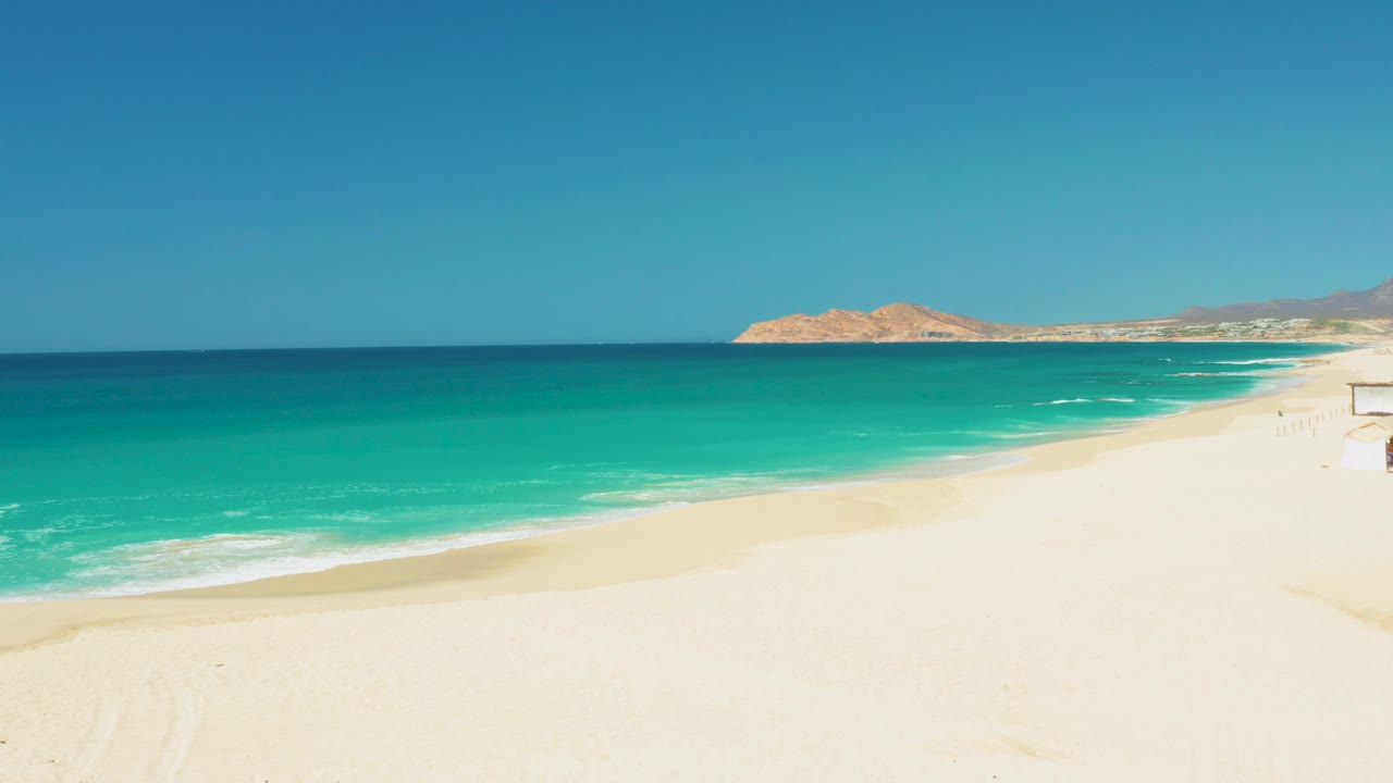 Dreams Los Cabos shoreline at 0:01 with the ocean band centered, white sand below, low tan hills in the distance, and no visible people.
