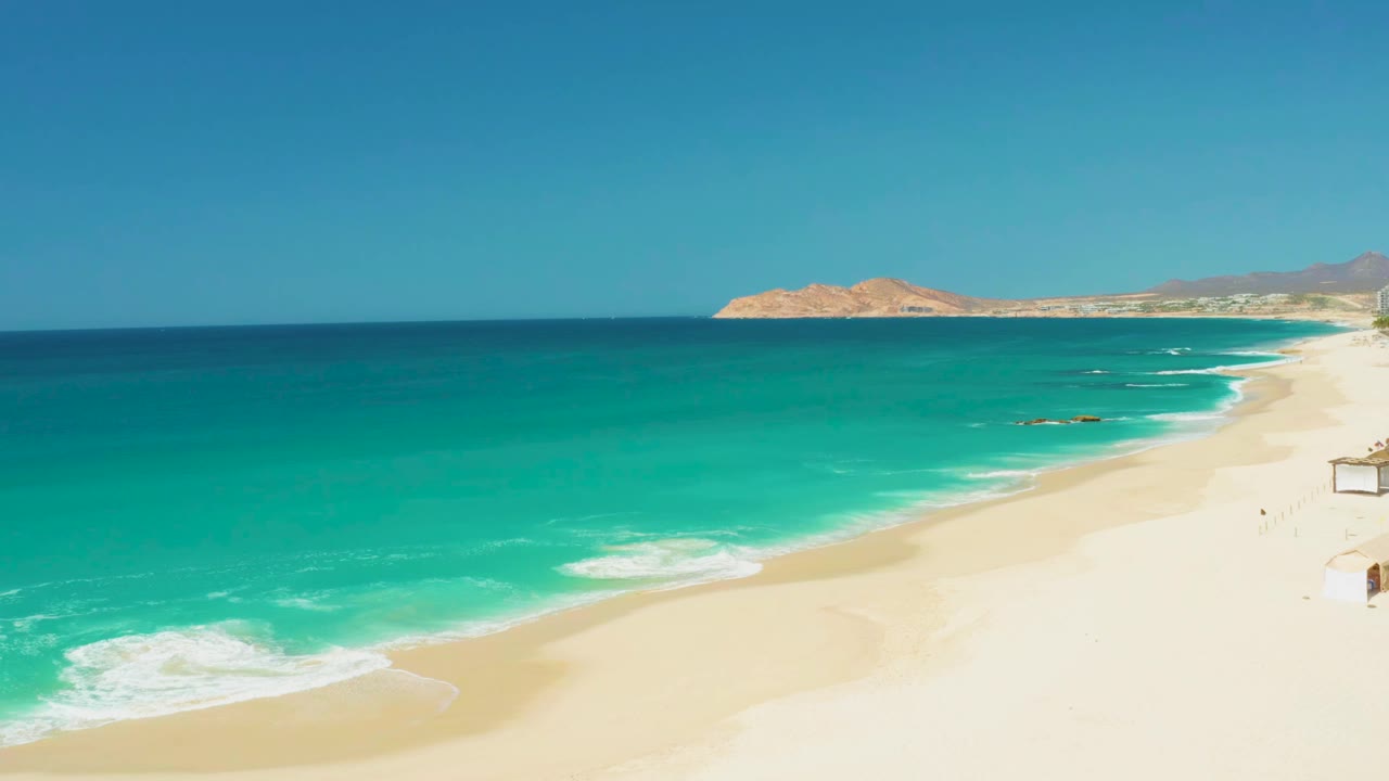Opening frame of the Dreams Los Cabos shoreline, with turquoise water on the left, pale sand on the right, distant landforms on the horizon, and no visible people.
