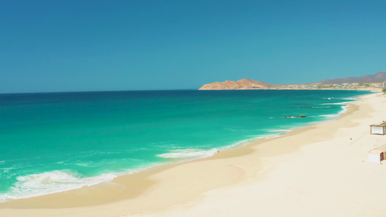 At 0:02, turquoise water fills more of the left side while white foam gathers near the lower shoreline at Dreams Los Cabos, with no visible people on the sand.