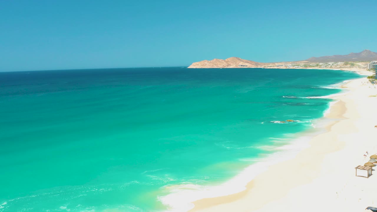 At 0:01, the Dreams Los Cabos shoreline angles through the frame as shallow turquoise water and white foam reach the sand, with no visible people.