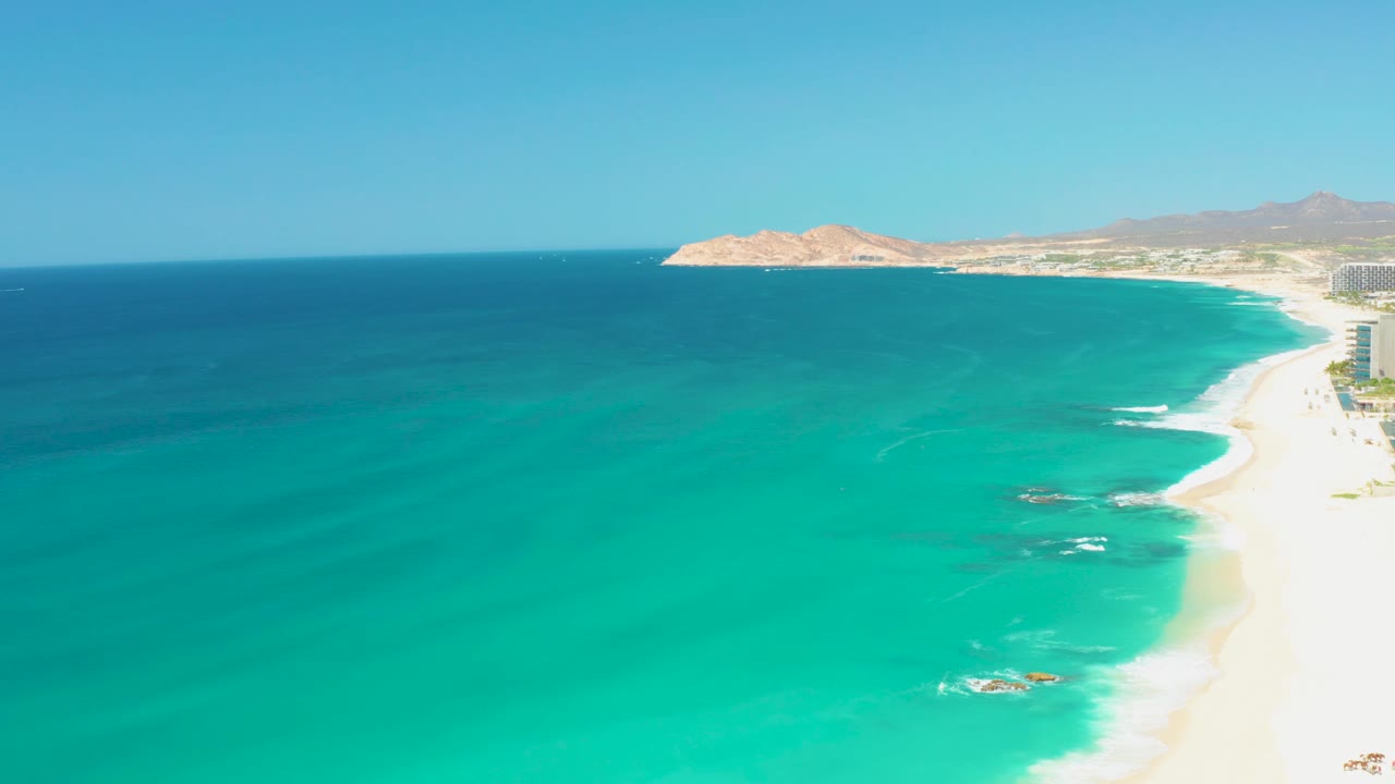 At 0:03, small white waves break around nearshore rocks below the Dreams Los Cabos coastline, with bright sand on the right and no visible people.