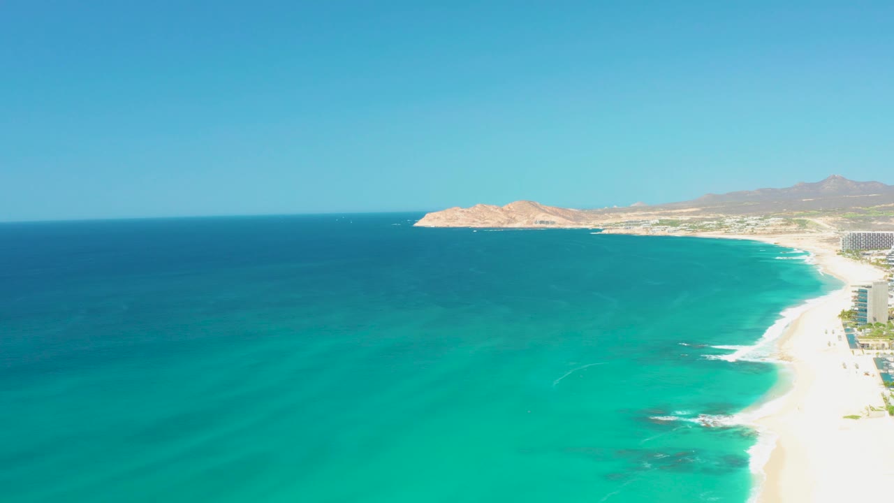 Aerial frame over Dreams Los Cabos with the surf line curling along the right side, deep blue water offshore, distant hills, and no visible people.