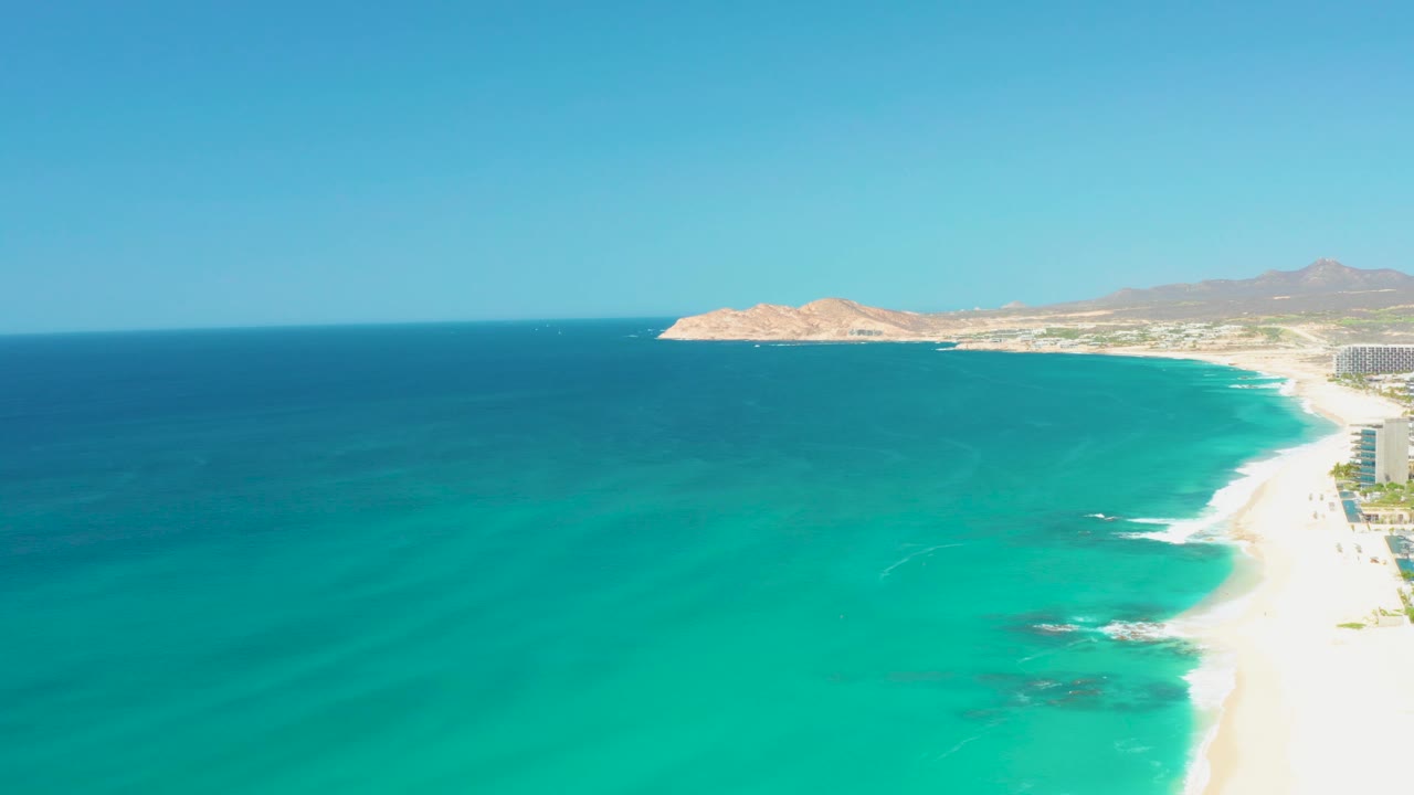 Mid-clip aerial frame at Dreams Los Cabos showing blue-green water, a bright sand strip at right, small foam breaks near shore, and no visible people.