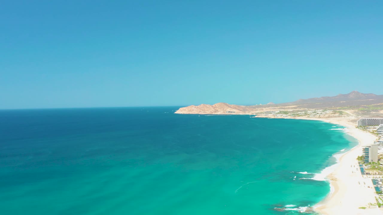 Aerial view at Dreams Los Cabos with the rocky point shifted toward the upper left, bright turquoise water across the center, a curved beach at right, distant buildings, mountains, and no visible people.