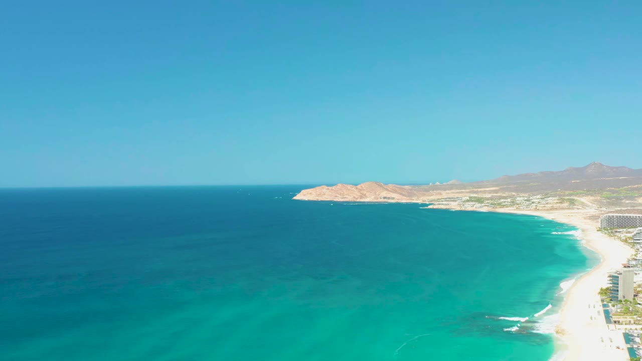 At 0:01, the Dreams Los Cabos coastline remains wide in frame, with deeper blue water to the left, white surf on the right-hand sand, distant mountains, and no visible people.