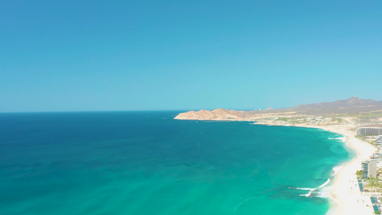At 0:02, the aerial view shows a large field of turquoise water, a narrow sandy beach with breaking waves, rocky landforms in the background, and no visible people.