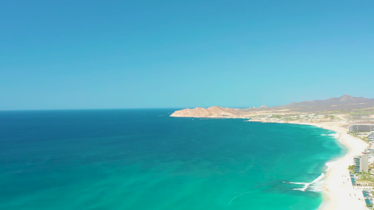 At 0:03, clear blue and turquoise water fills the foreground, small white waves touch the sandy shore at right, mountains sit beyond the coast, and no visible people appear.