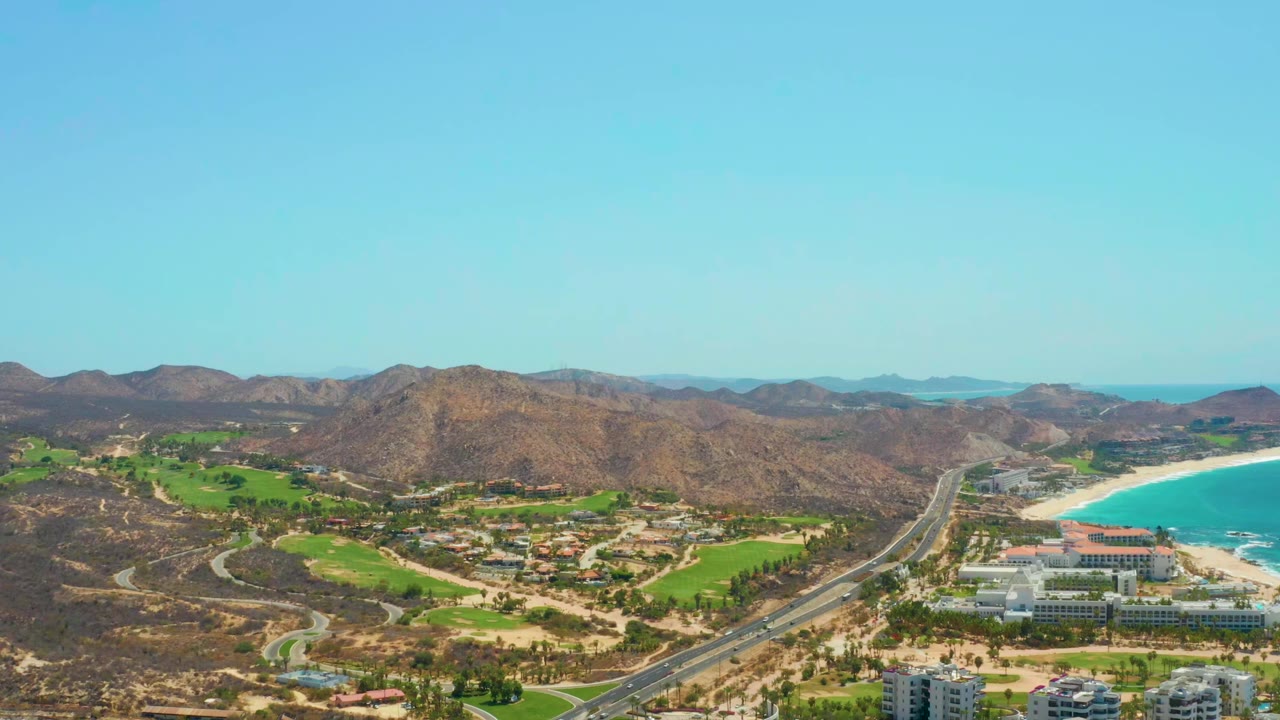 At 0:01, the Dreams Los Cabos aerial keeps the highway centered between green landscaped grounds and the beach, with resort-area buildings near the shore, dry hills in back, and no visible people.