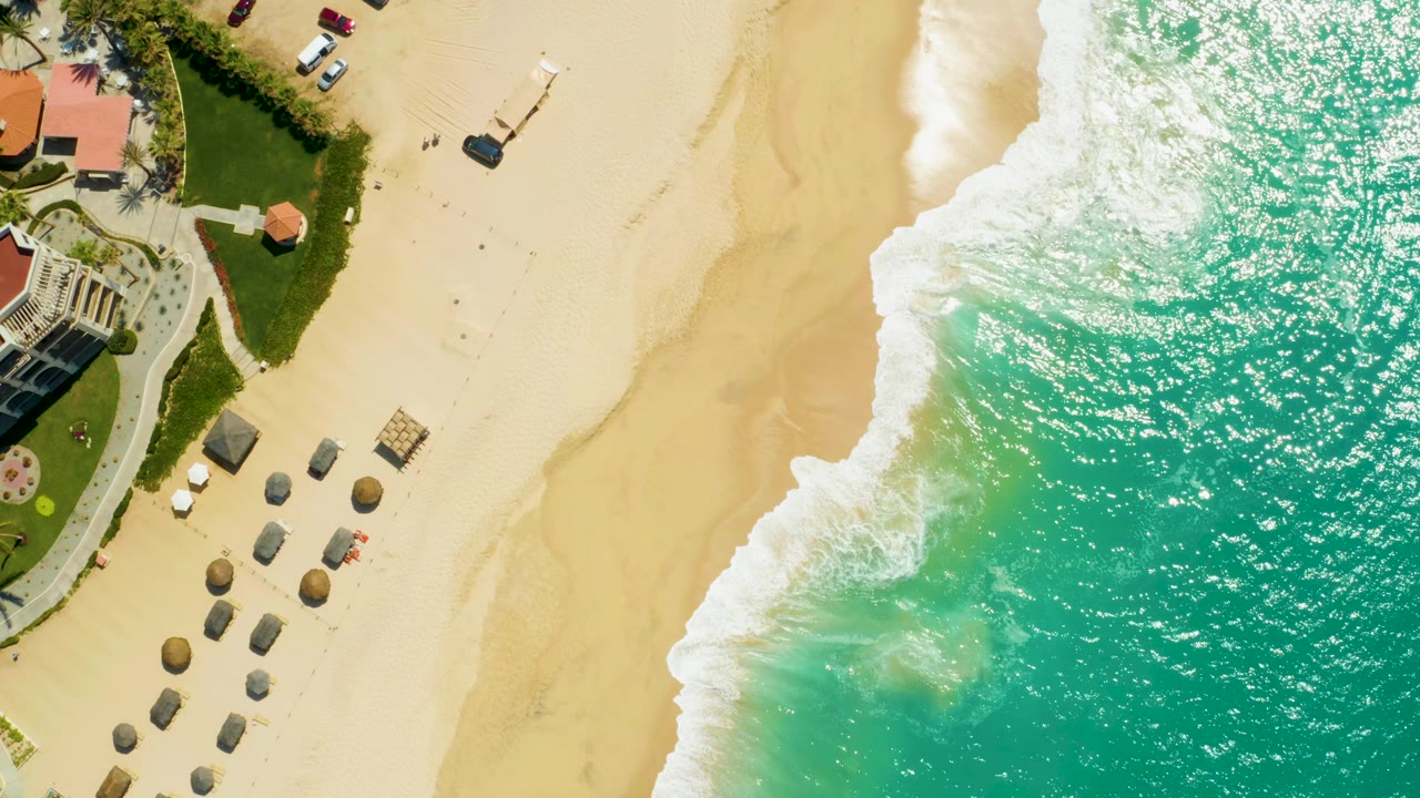 Top-down aerial frame at Dreams Los Cabos shows turquoise surf on the right, pale sand in the center, a row of umbrellas or shade huts near the lower left, resort landscaping, and no visible people.