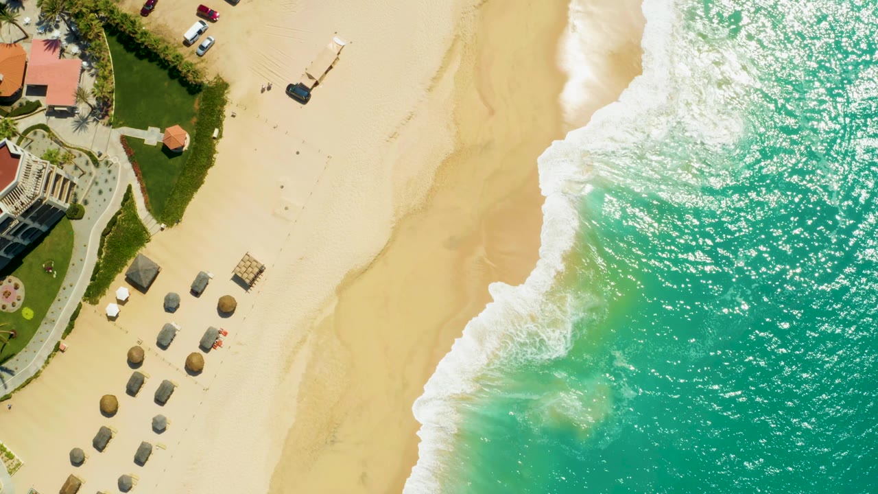 Aerial beach frame with waves crossing sand beside resort landscaping.