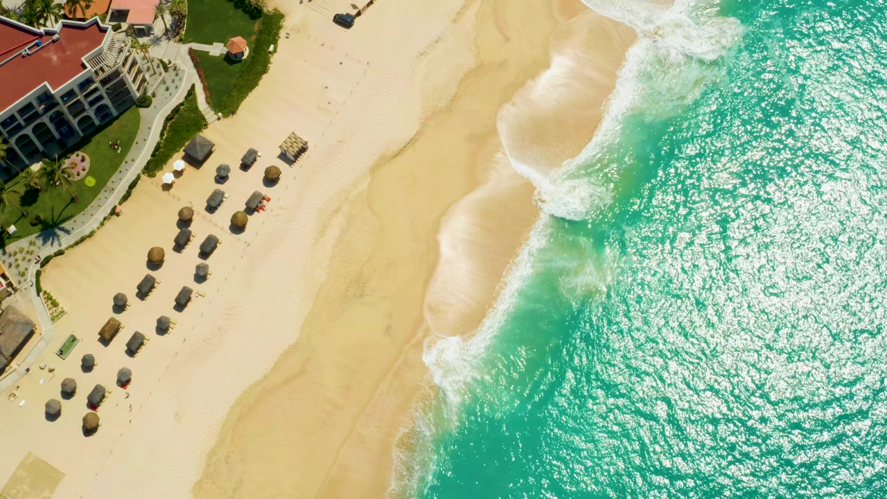 At 0:01, the aerial view holds the Dreams Los Cabos building at upper left while foamy waves meet the open sand beside rows of shade umbrellas, with no visible people.