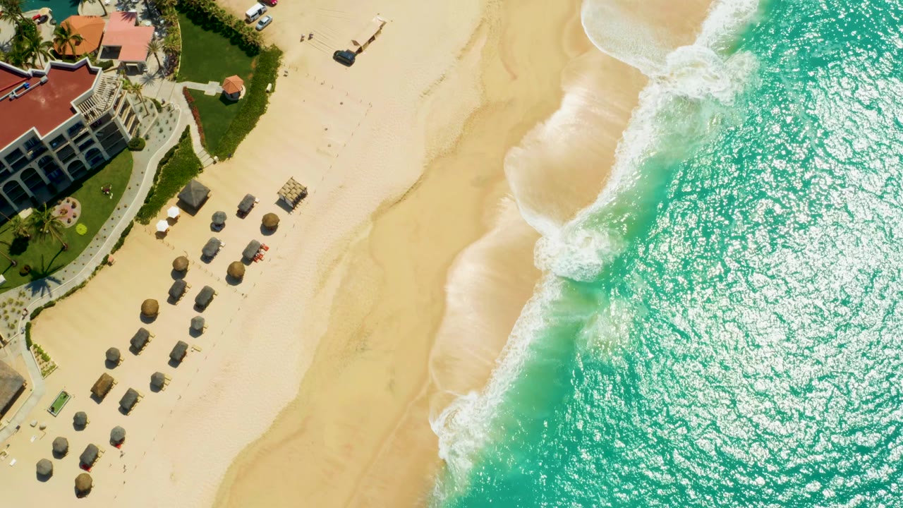 At 0:02, the frame shifts slightly along the shoreline, showing more open sand between the Dreams Los Cabos resort grounds and the bright turquoise ocean, with no visible people.