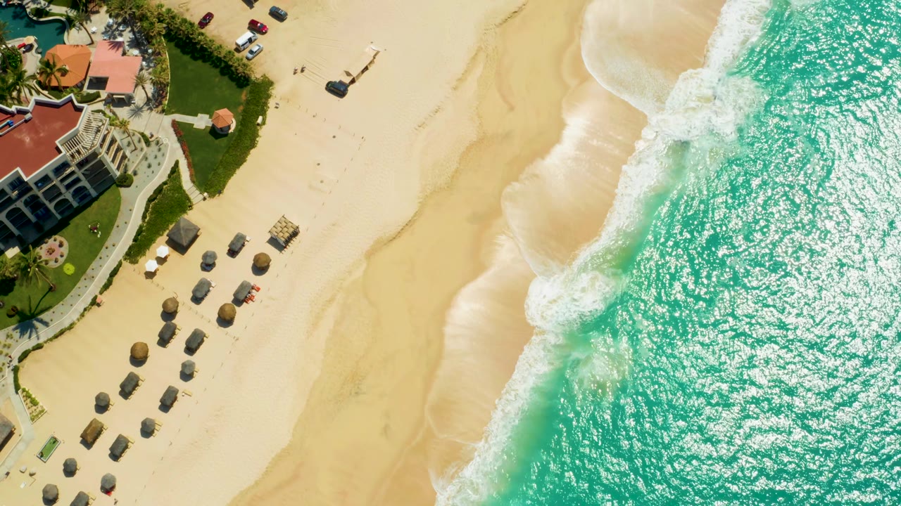 At 0:03, the resort edge sits near the left border while the shoreline curves through the frame and sunlit turquoise water fills the right side, with no visible people.