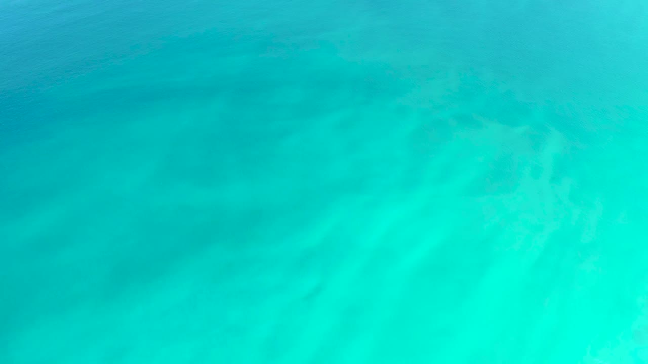 Clear blue-green ocean water reveals soft sand markings and a darker underwater area, with no visible people.