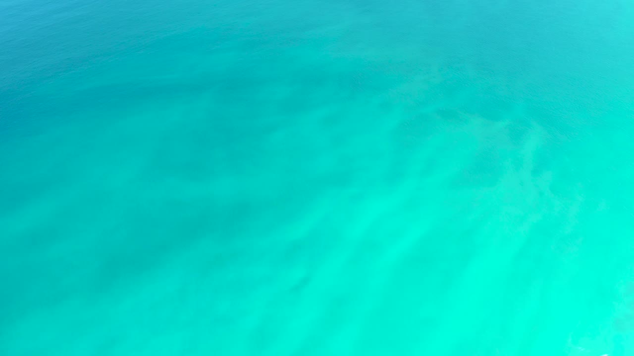 Shallow turquoise sea covers a pale sandy floor, with a darker patch near the upper left and no visible people.