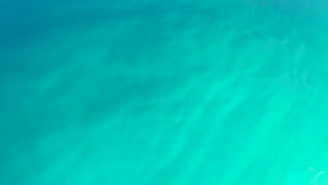 Bright turquoise shallows at Dreams Los Cabos fill the frame above pale sand, with faint surface texture and no visible people.