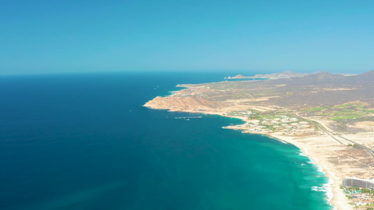 Opening aerial frame over Dreams Los Cabos in Baja California Sur, Mexico, with deep blue water at left, turquoise surf along the beach, a coastal road inland, distant hills, and no visible people.