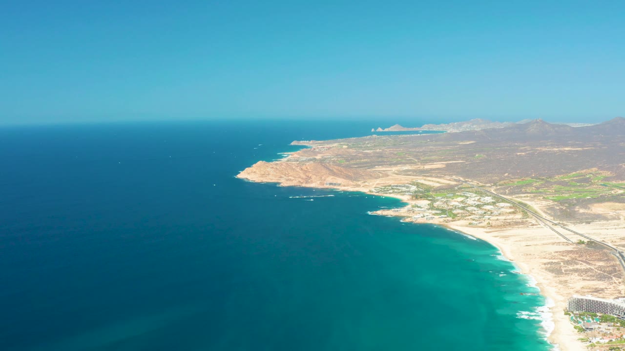 Frame at 0:02 shows Dreams Los Cabos coastline from above, with darker water offshore, brighter turquoise water near the beach, a road through dry land, a resort edge at lower right, and no visible people.