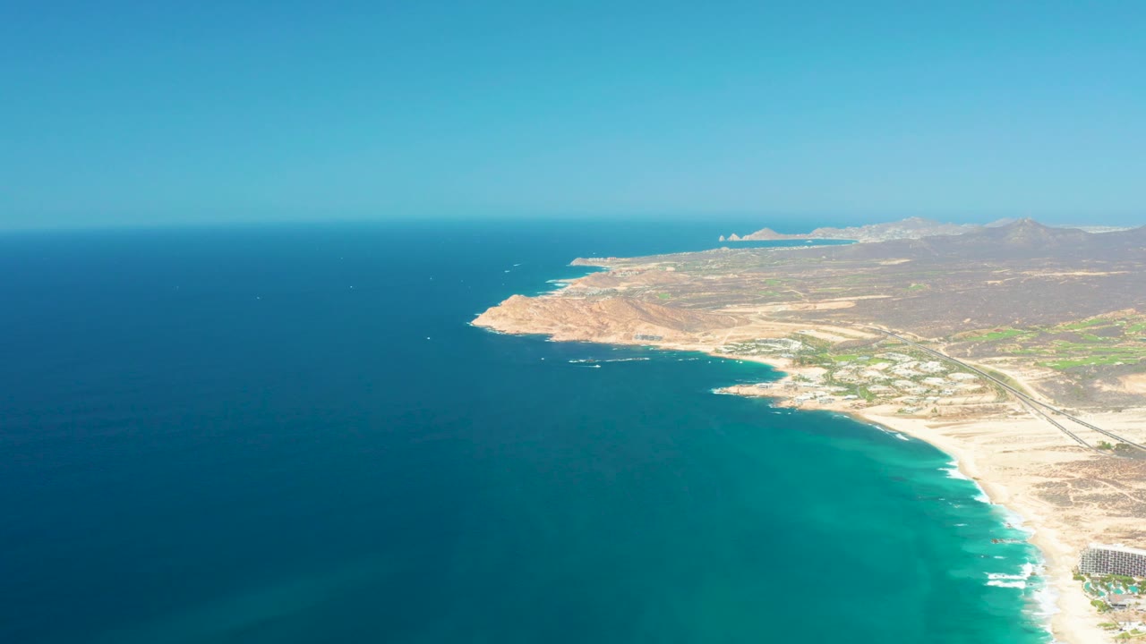 High aerial frame of the Dreams Los Cabos coastline, with deep blue water offshore, lighter turquoise water near the sand, dry hills in the distance, and no visible people.