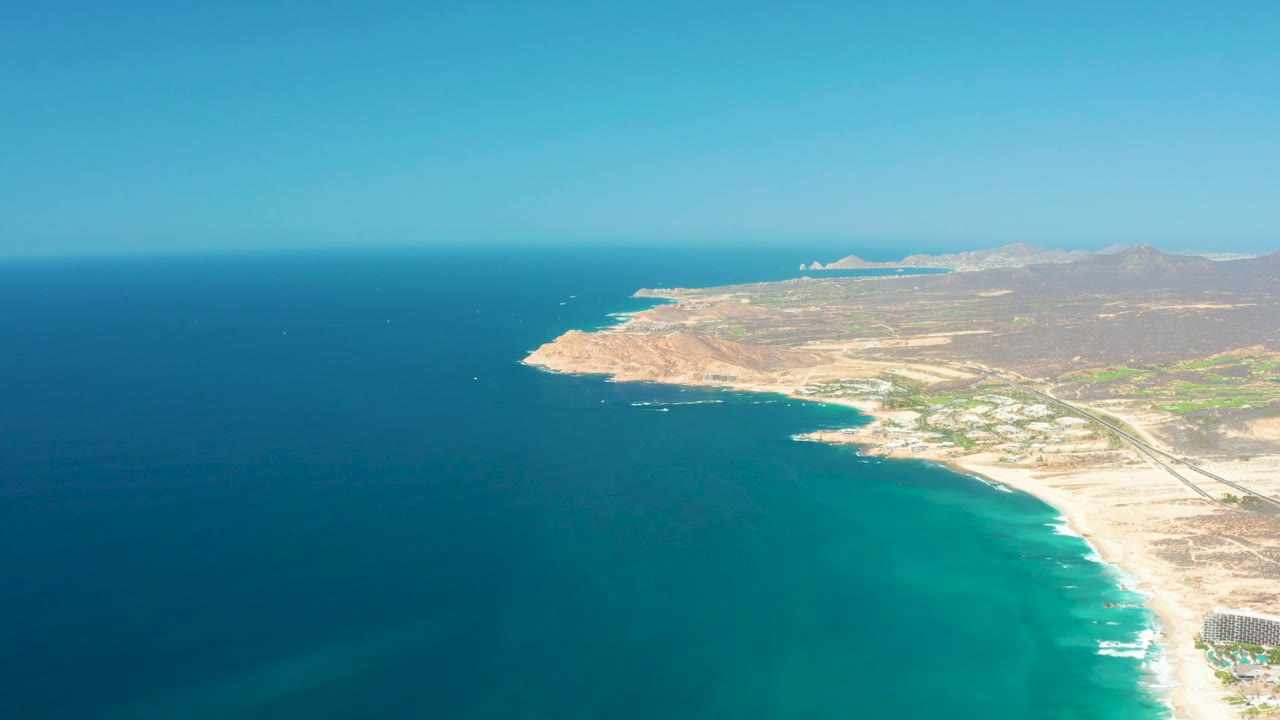 Aerial view of Dreams Los Cabos with a curved sandy shoreline, calm blue-green water, pale inland terrain, distant hills, and no visible people in the frame.