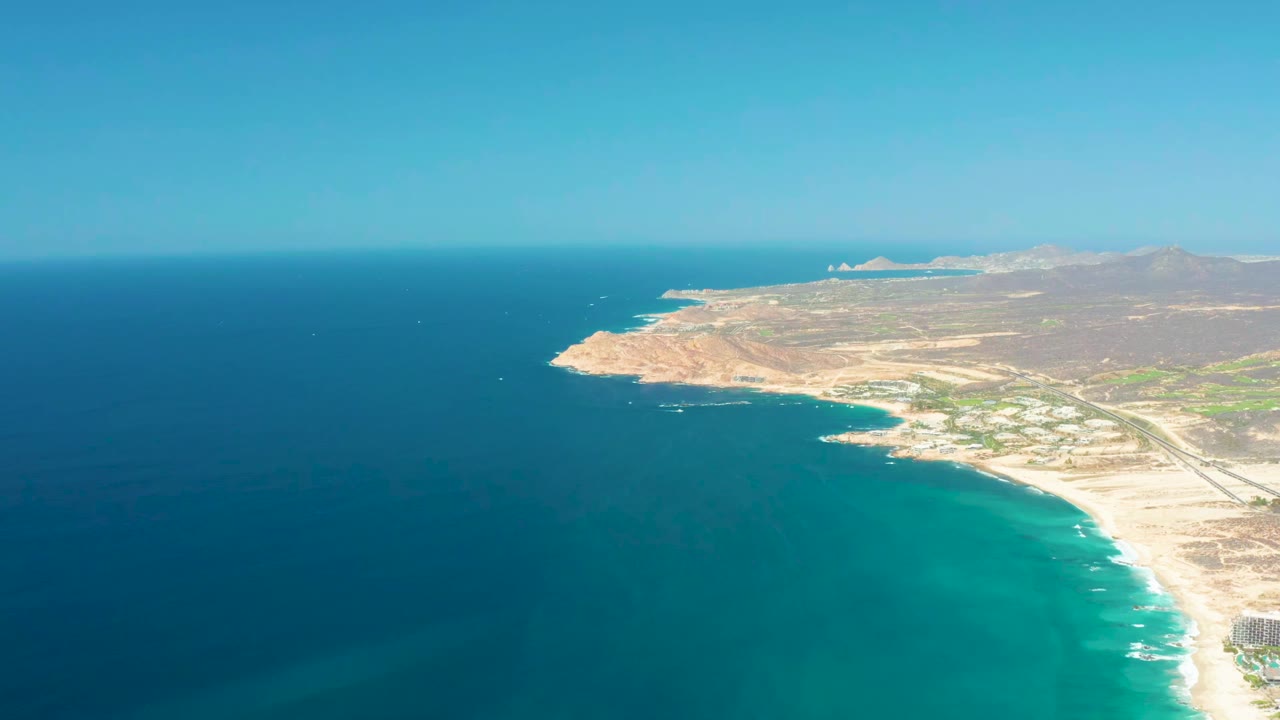 Aerial frame over Dreams Los Cabos showing the coast slightly farther along the bay, with turquoise water at the beach edge, deep ocean offshore, dry hills beyond, and no visible people.
