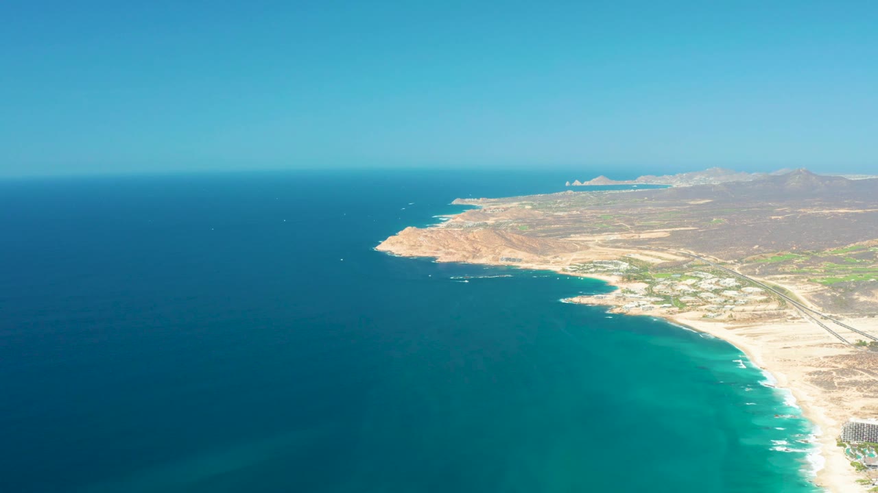 Aerial coast scene with blue water, sandy shoreline, and hills under clear sky.