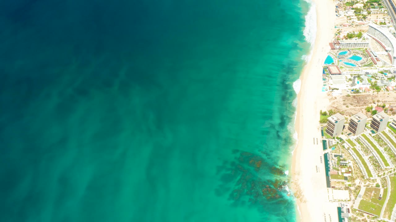 Aerial frame at Dreams Los Cabos with broad teal water, darker underwater patches near shore, a narrow white beach, resort pools, buildings, and no visible people.
