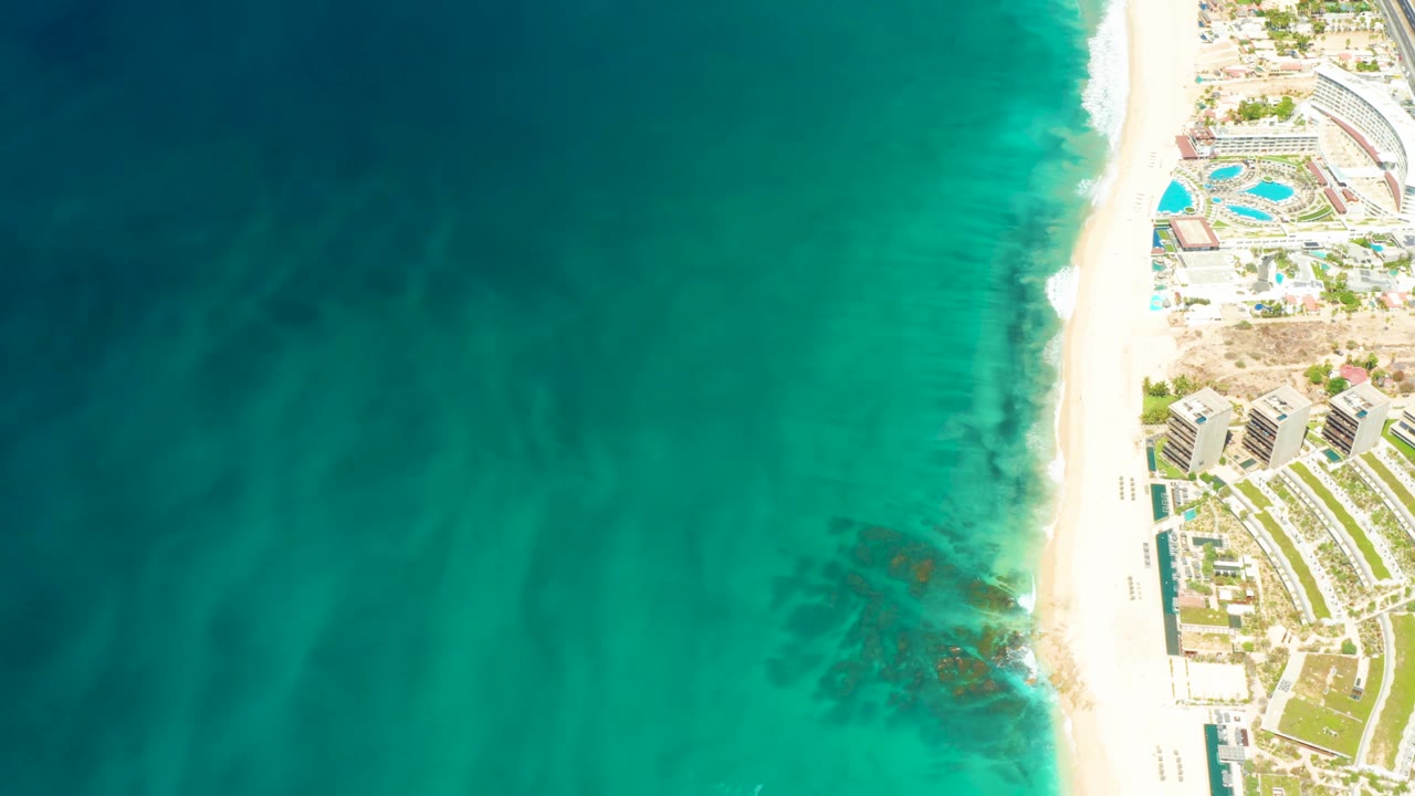 High overhead view at Dreams Los Cabos showing turquoise water, pale beach, curved blue pools, resort buildings, planted grounds, and no visible people.
