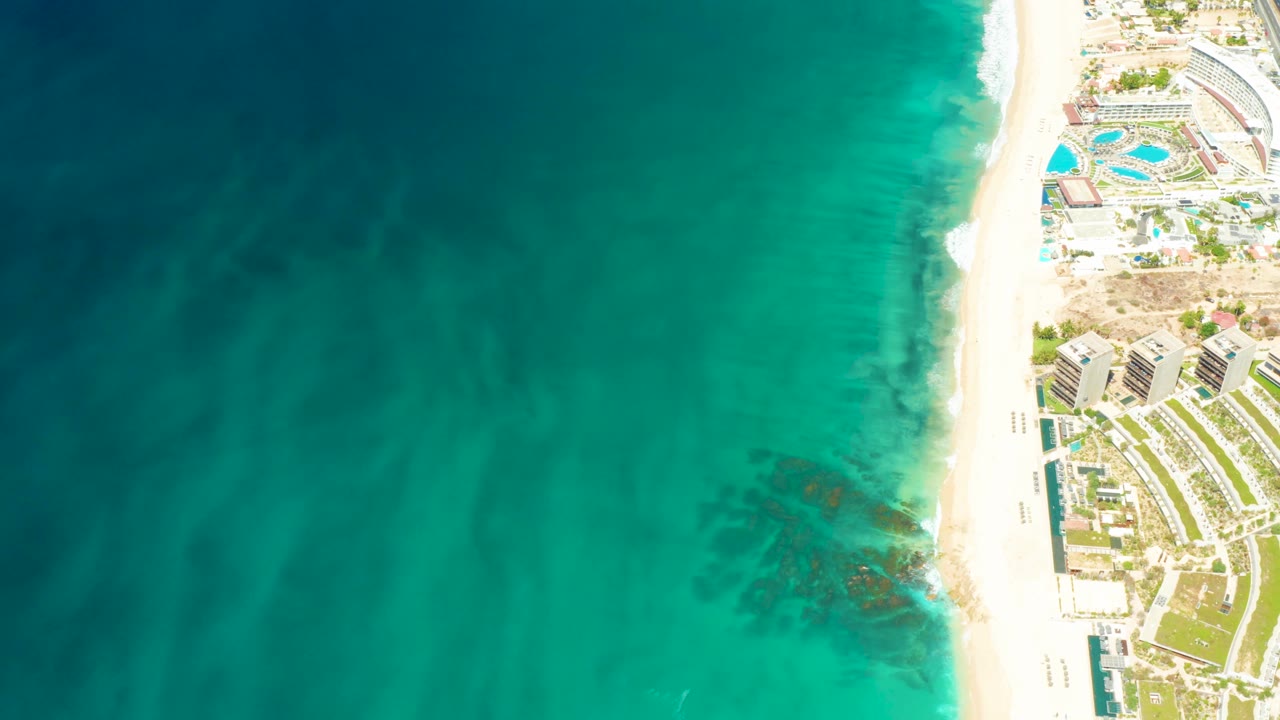 Overhead Dreams Los Cabos shoreline view with subtle wave texture in turquoise water, white sand along the right edge, resort buildings and pools beyond it, and no visible people.