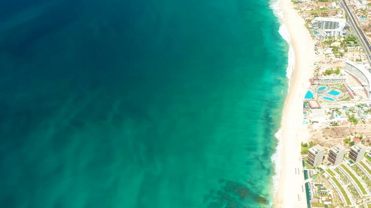 Aerial frame over Dreams Los Cabos showing clear green-blue water, a narrow white beach, resort pools, buildings, lawns, and a road near the top right, no visible people.