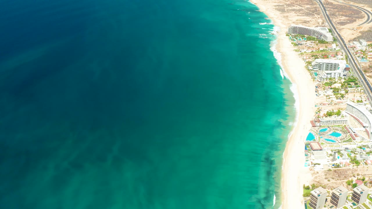 Aerial frame over Dreams Los Cabos showing deep teal water, pale surf, sandy beach, resort buildings, pools, and no visible people at the start of the clip.