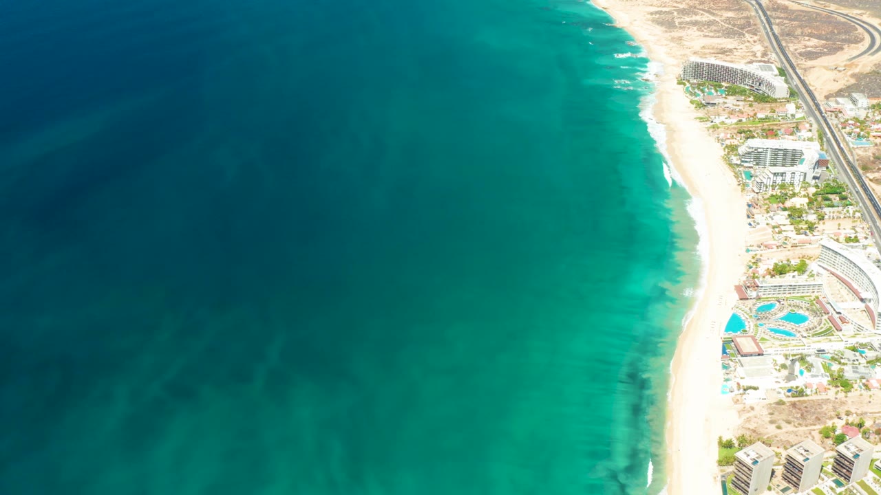 Aerial view over Dreams Los Cabos with broad teal ocean, a narrow surf line, bright sand, resort pools, and no visible people one second into the shot.