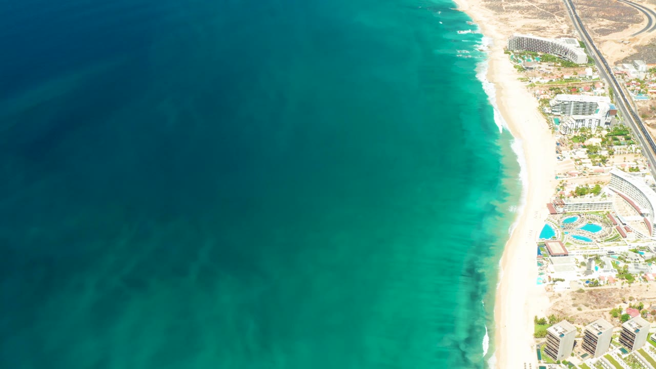 Drone frame over Dreams Los Cabos showing darker ocean patches offshore, foamy waves at the sand, resort buildings near pools, and no visible people.