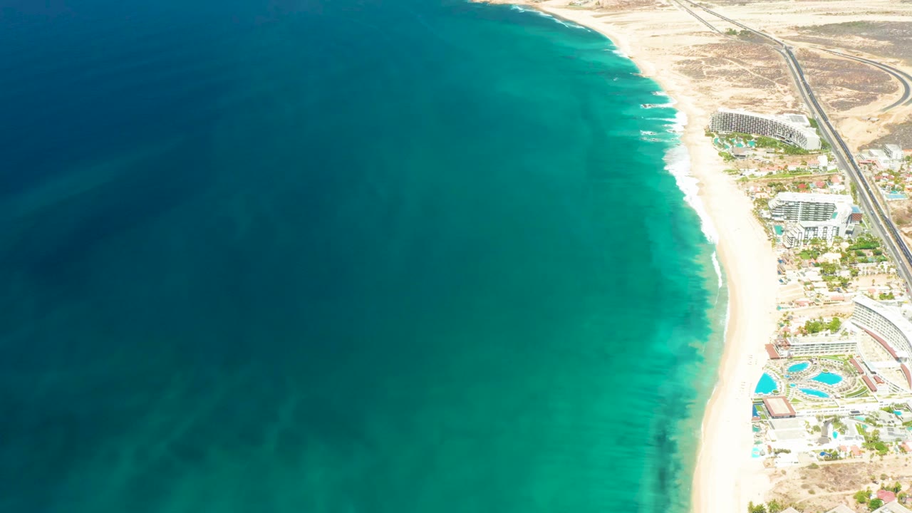 High aerial frame over Dreams Los Cabos showing deep blue water shifting to turquoise shallows, a pale beach, resort buildings, pools, road, and no visible people.