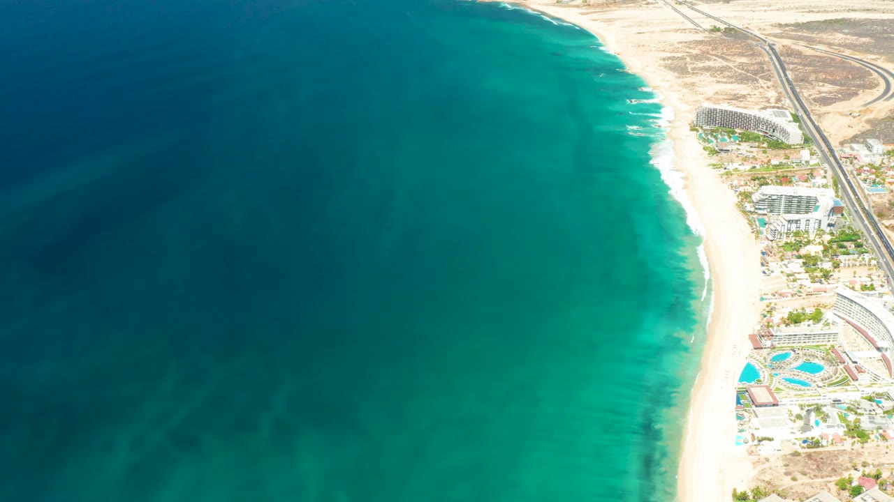 High aerial frame over Dreams Los Cabos where darker offshore water fills most of the image and the resort strip, road, pools, sand, and no visible people sit along the right edge.