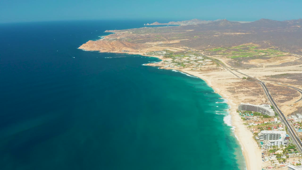 Opening aerial view over Dreams Los Cabos with turquoise ocean on the left, a sandy beach and parallel road on the right, arid hills in the distance, and no visible people.