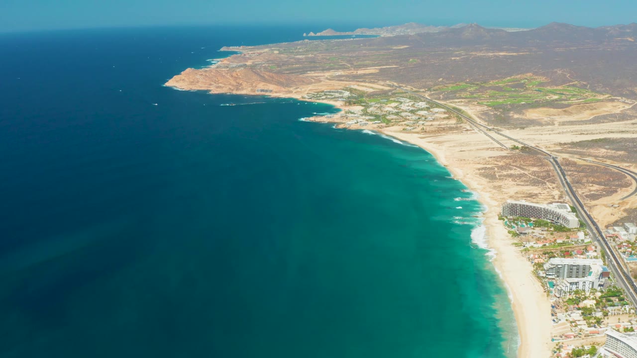 The frame shows Dreams Los Cabos shoreline from above, with broad turquoise water, pale beach, resort buildings near the lower right, a road, arid hills, and no visible people.