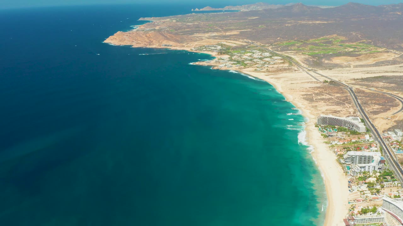 Mid-clip aerial view over Dreams Los Cabos with deep blue water in the left foreground, turquoise shallows along the beach, roadside buildings, dry hills, and no visible people.