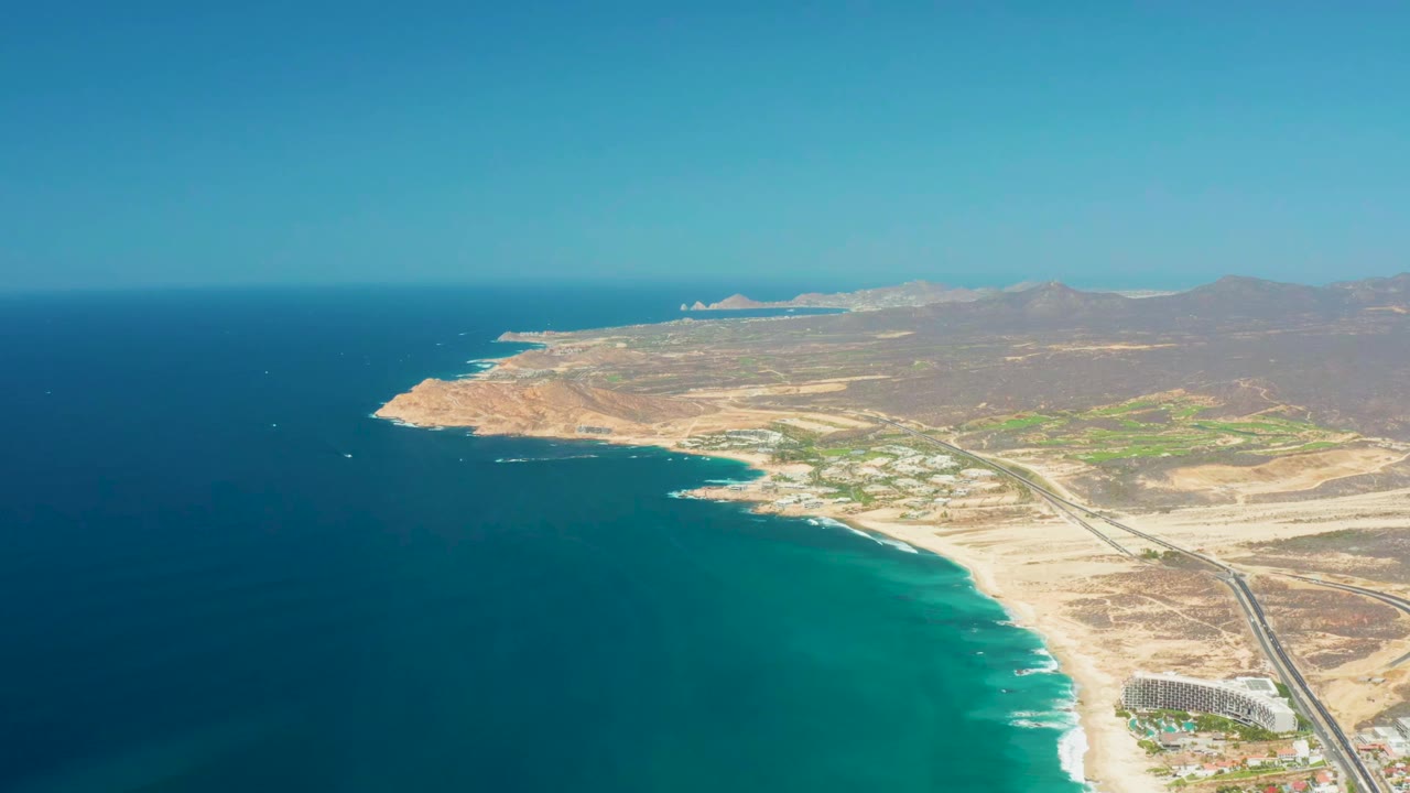High aerial beach view with turquoise water, white surf, desert roads, and mountain horizon.